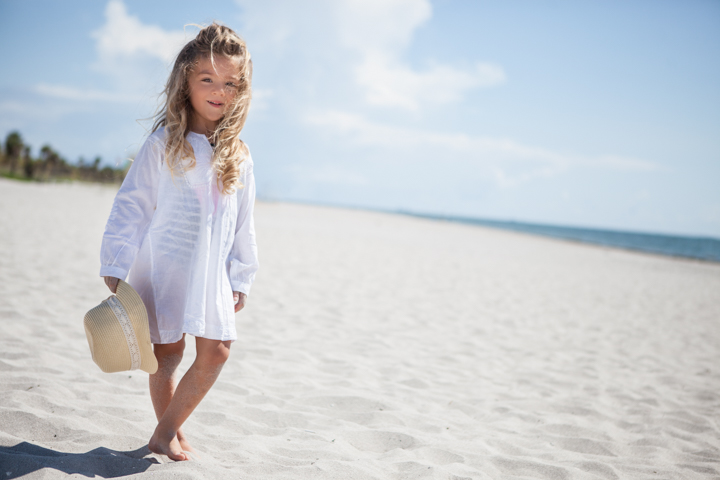 A child in a white dress carries a small bag across a sandy beach.