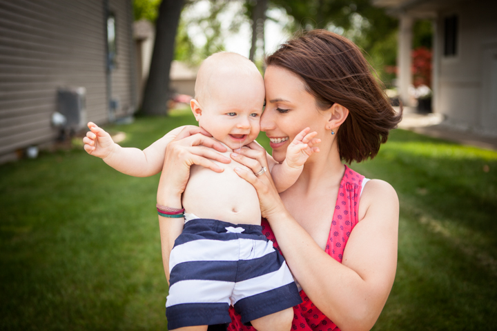 A smiling woman holds a baby close on a sunny lawn.