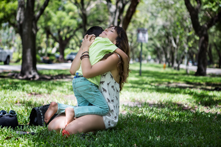 A woman kneels on grass and hugs a child in a green park.
