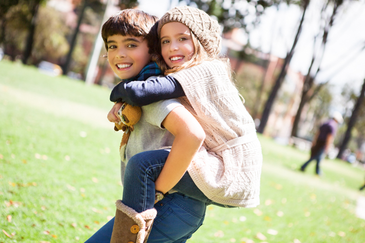 A smiling child carries another child on their back in a sunny park.