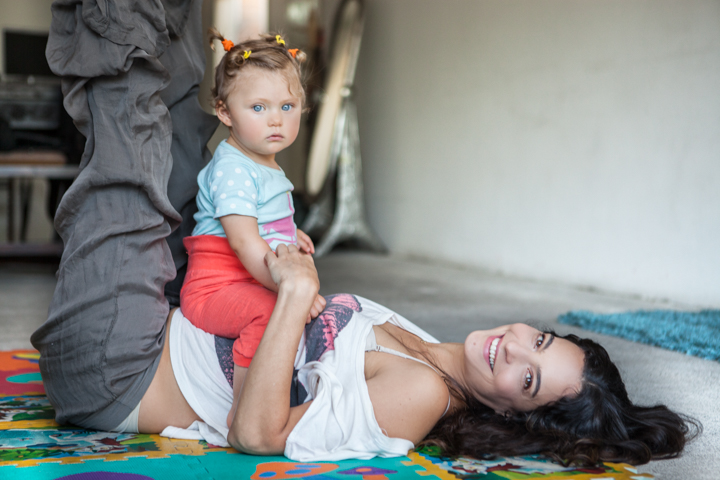 A woman balances a toddler on her raised legs while lying on a rug.