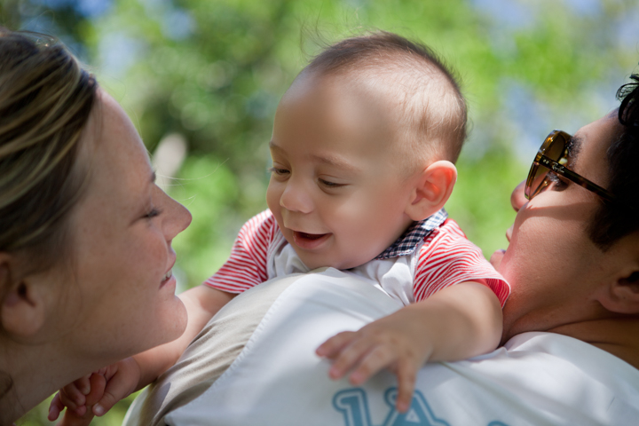 A smiling baby is held between two adults in a green garden.