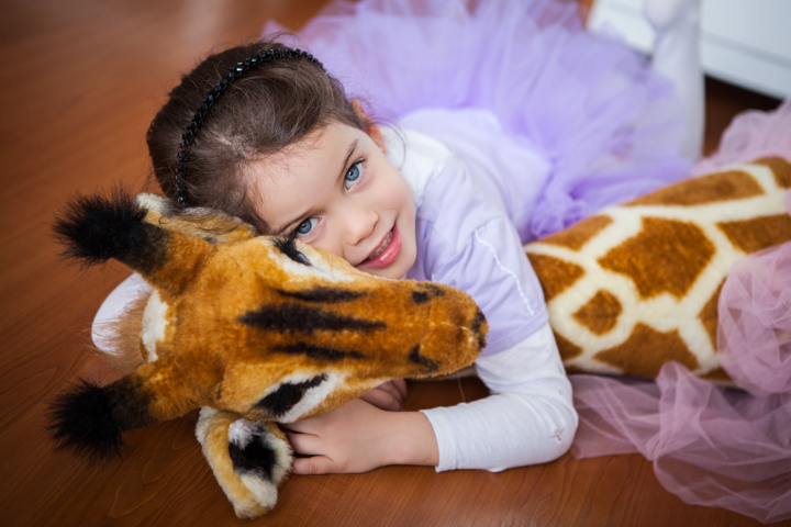 A laughing child hugs a large stuffed giraffe on a wooden floor.