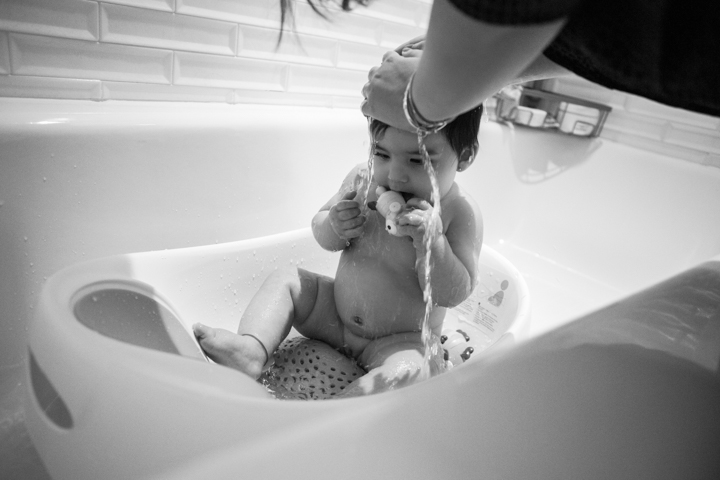 A baby sits in a bathtub holding an adult's hand in black and white.