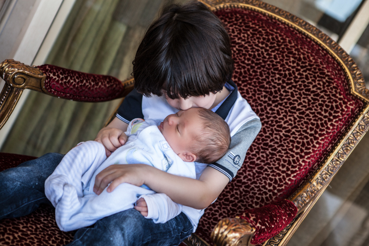 A child cuddles a newborn while sitting in an upholstered chair.