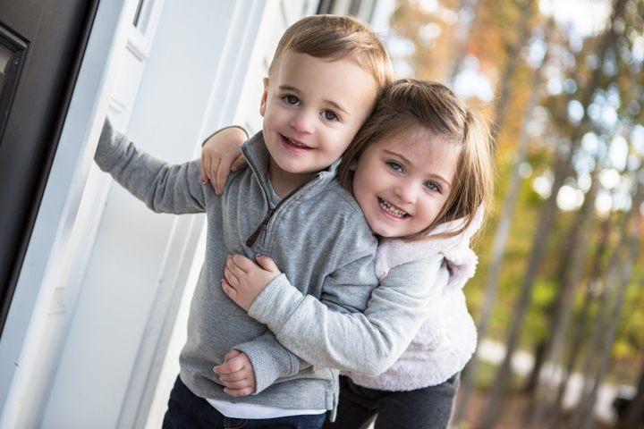 Two smiling children hug beside a bright window.