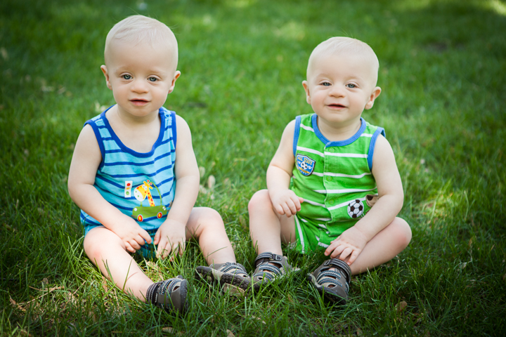Two babies in bright striped outfits sit side by side on grass.