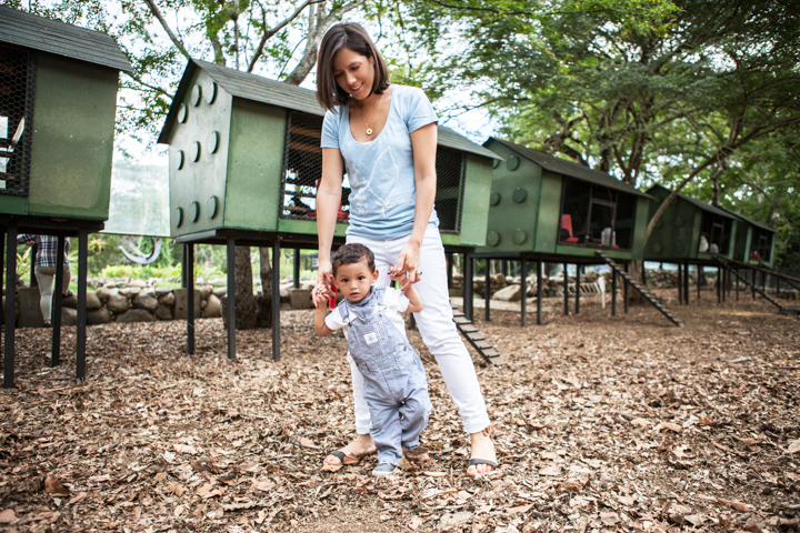 A woman steadies a toddler walking across a woodchip playground.