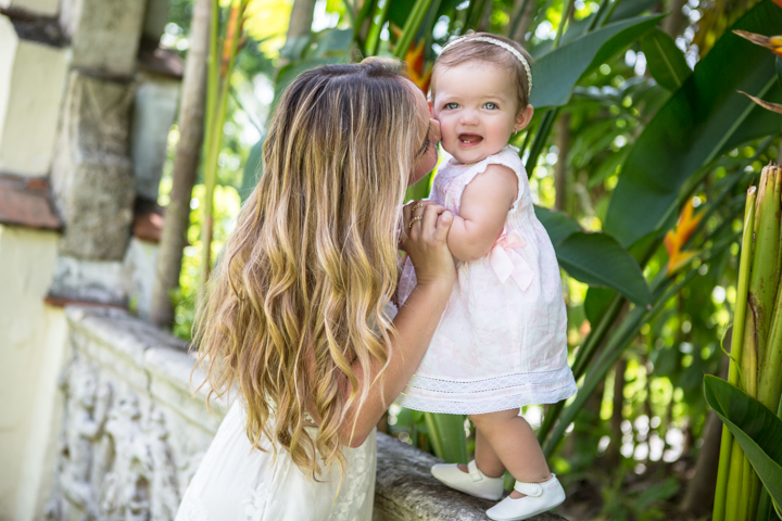 A woman lifts a smiling toddler beside a leafy tree.