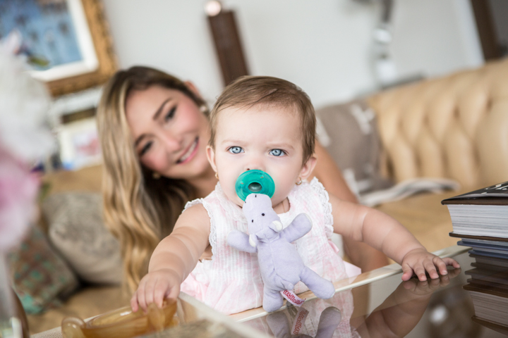 A baby with a green pacifier leans on a table while a woman smiles behind them.