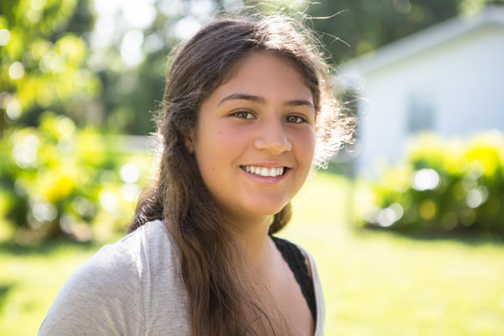 A smiling child with long dark hair stands on a sunny lawn.