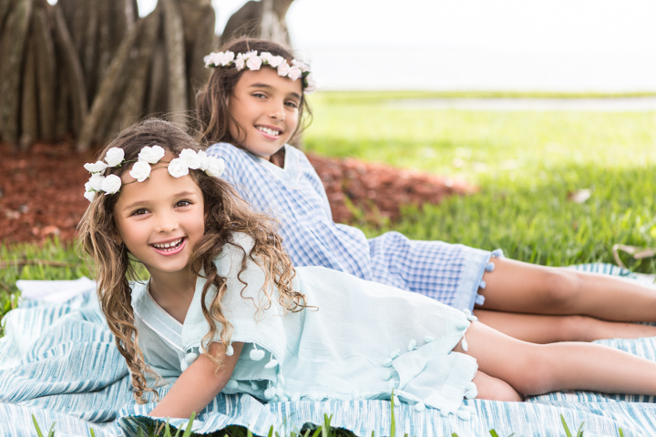 Two children wearing flower crowns relax on a blanket beneath a tree.