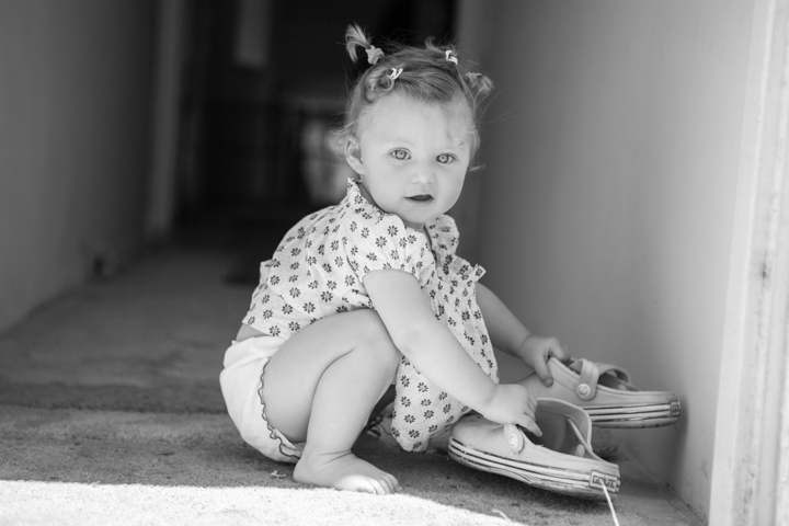 A toddler crouches beside oversized shoes in a black-and-white photograph.