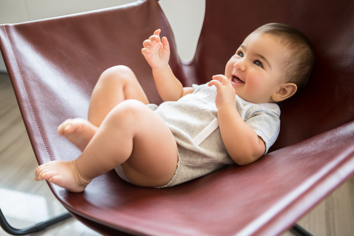 A smiling baby reclines in a curved chair with feet raised.