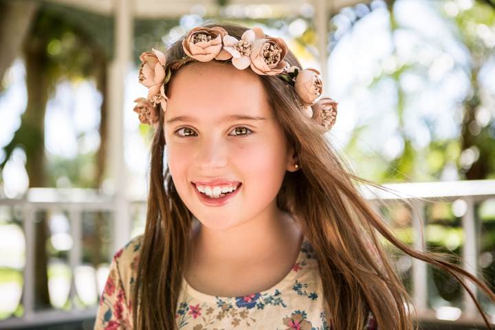 A smiling child wears a pink flower crown beneath green trees.