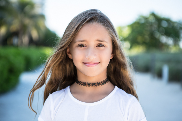 A smiling child in white wears a dark choker outdoors.