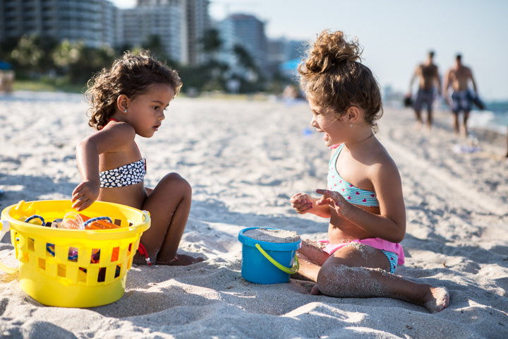 Two children play with buckets in the sand on a sunny beach.