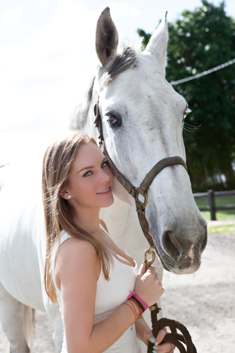 A woman in white rests beside a white horse outdoors.