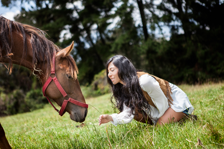 A woman kneels in the grass and reaches toward a brown horse.