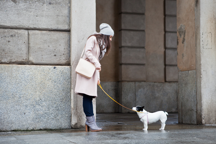 A woman in a pale coat walks a small white dog beside a stone building.