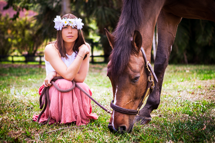 A woman wearing a flower crown sits in the grass beside a grazing horse.