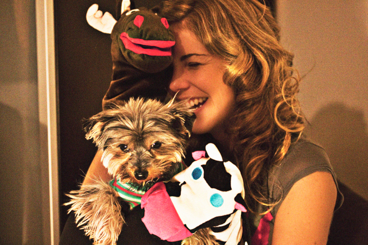 A smiling woman holds a small dog dressed in a colorful costume.