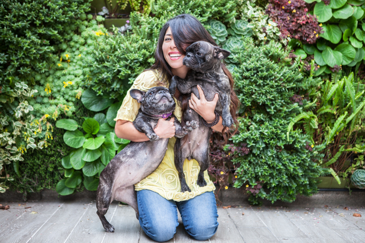 A woman kneels with two small dogs beside a leafy green wall.