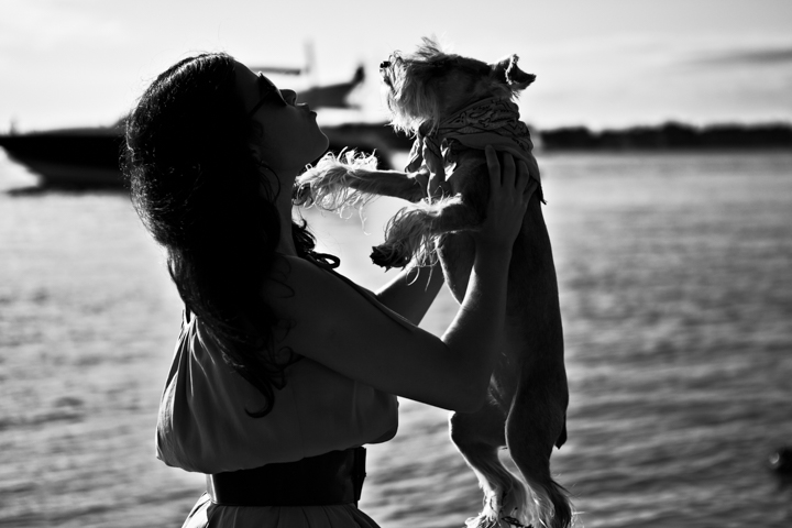 A woman lifts a dog beside the water in a black-and-white silhouette.