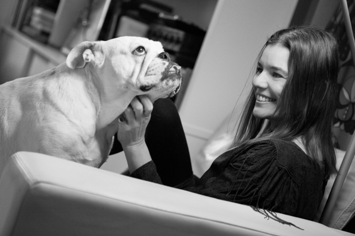 A smiling woman holds a small dog while reclining in a black-and-white portrait.