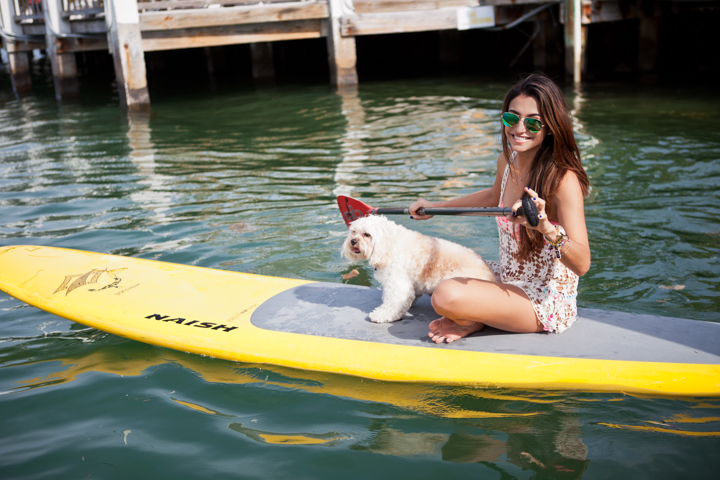 A woman sits on a yellow paddleboard with a small dog on calm water.