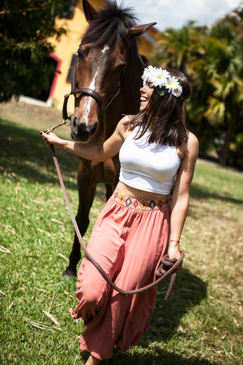 A woman in a white top and pink skirt stands beside a horse in a sunny pasture.