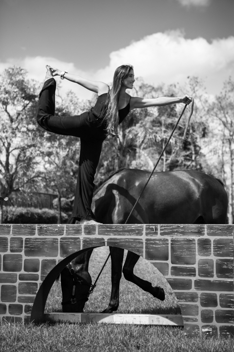 A person raises an arm while standing beside a horse in black and white.