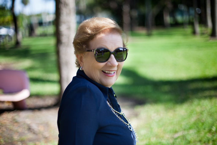 A smiling woman in sunglasses turns toward the camera in a green park.