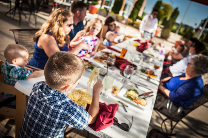 Adults and children share a meal around a long outdoor table.