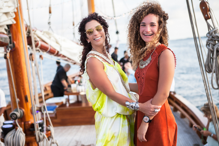 Two women in colorful dresses stand together aboard a sailboat.