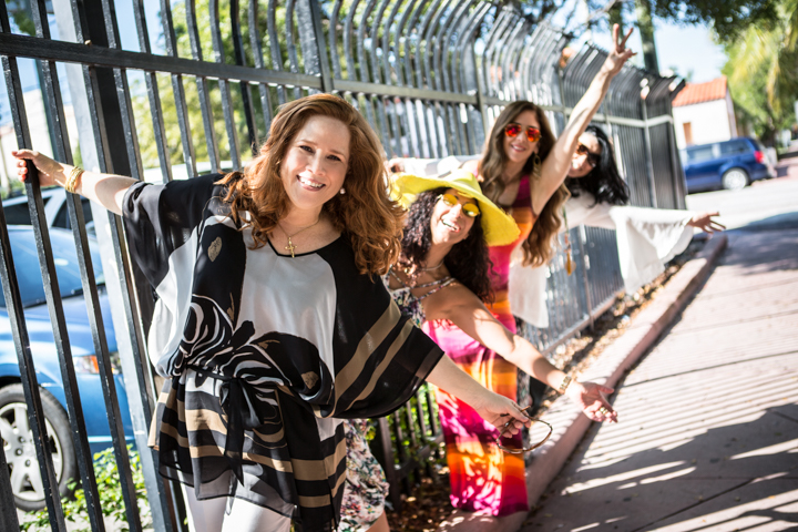 Three women smile along a waterfront railing on a sunny walkway.