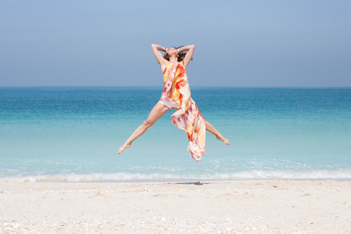 A woman in a colorful dress jumps above the sand beside turquoise water.