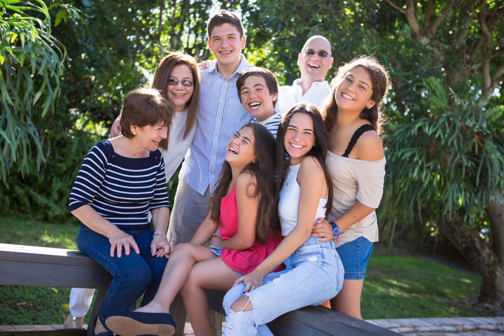 Adults and children gather closely on a bench beneath green trees.