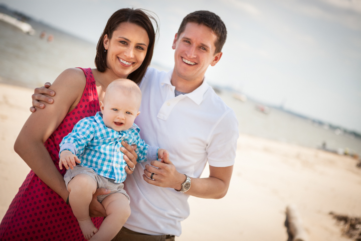 Two smiling adults hold a baby together on a sandy beach.