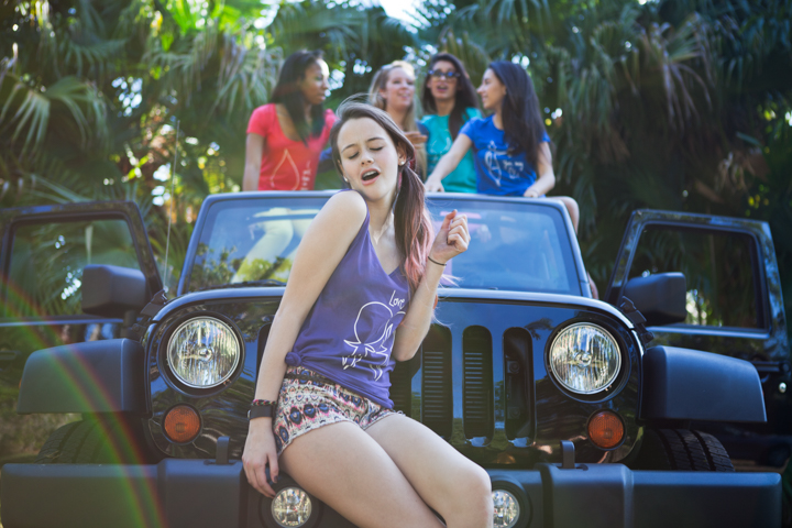 A group of women gathers around an open-top vehicle beneath tropical plants.