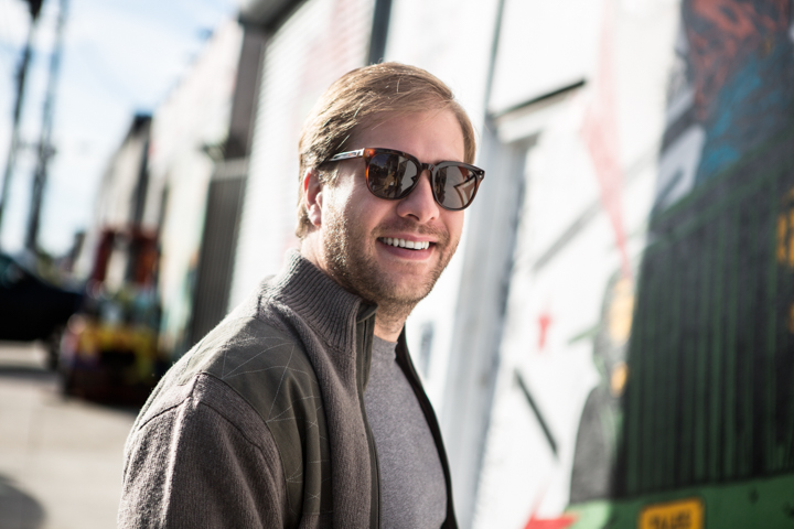 A smiling man in sunglasses stands on a city street.