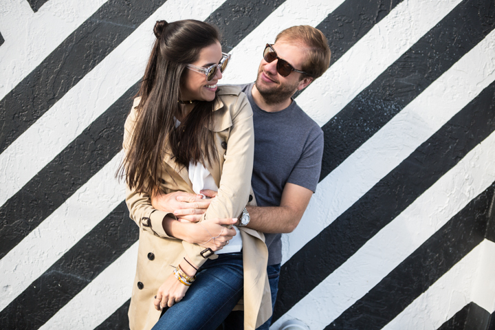 Two adults smile at each other while holding a young child beside a striped wall.