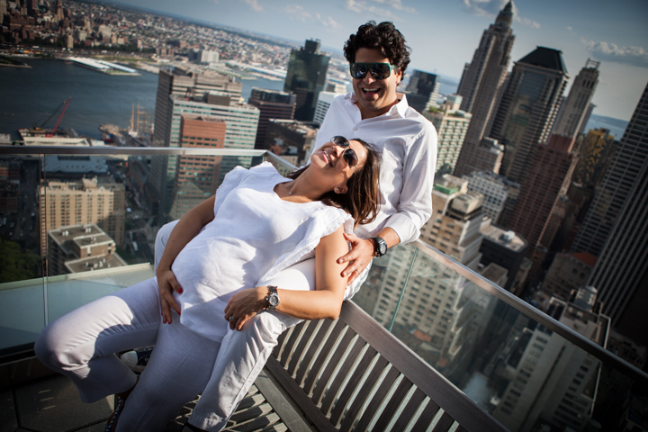 Three adults laugh together on a balcony overlooking a city skyline.