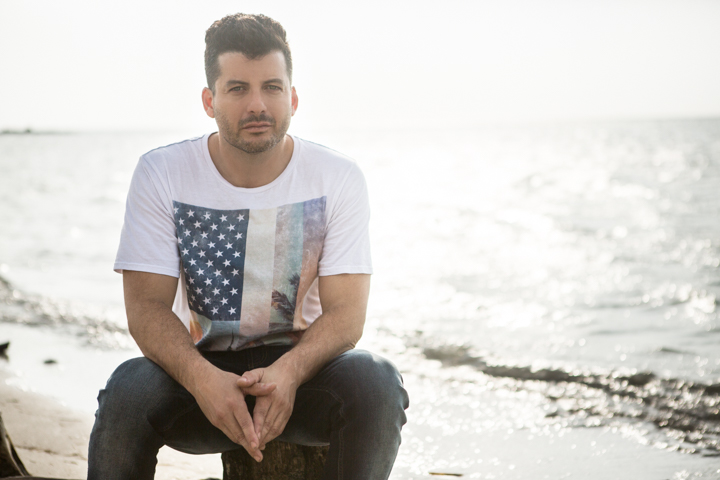 A man in a printed T-shirt sits beside sunlit ocean water.
