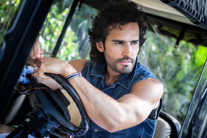 A man in a sleeveless denim shirt leans on the steering wheel of an open vehicle.