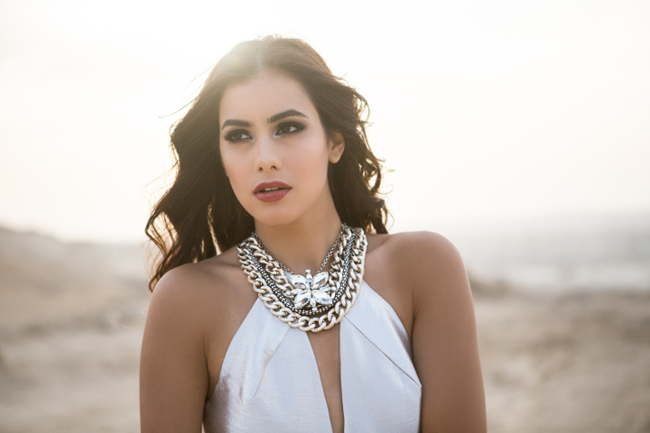 A woman in white wears layered silver necklaces in warm outdoor light.