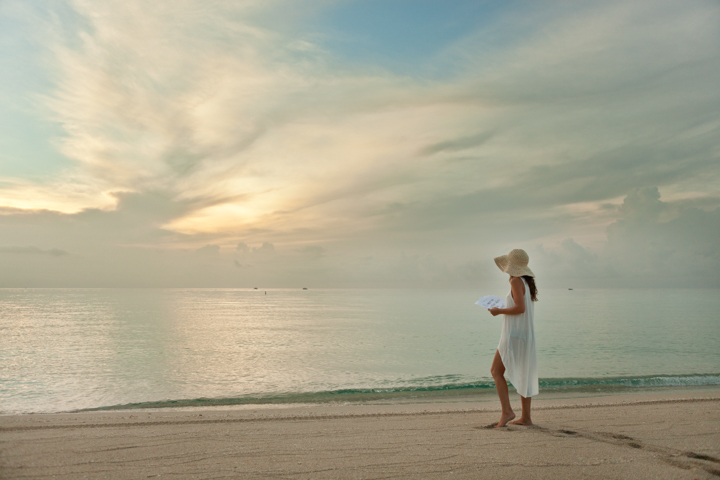A person wearing a hat stands on a beach beneath a cloudy sunset sky.