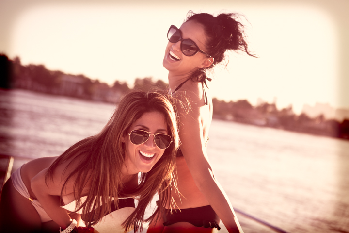 Two women in sunglasses smile together in warm seaside light.