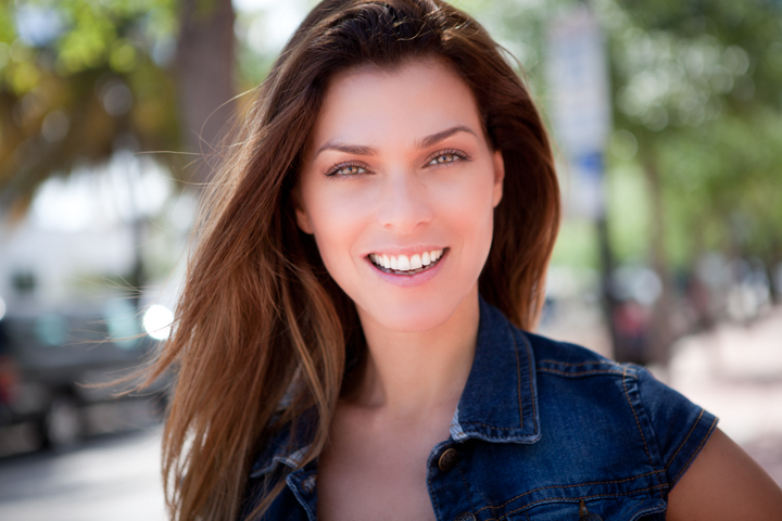 A smiling woman with long brown hair wears a sleeveless denim top outdoors.