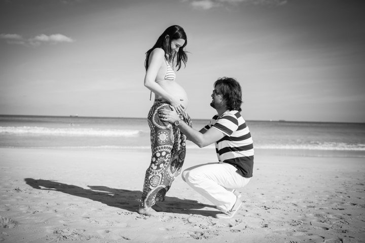 A man kneels beside a pregnant woman on a beach in black and white.